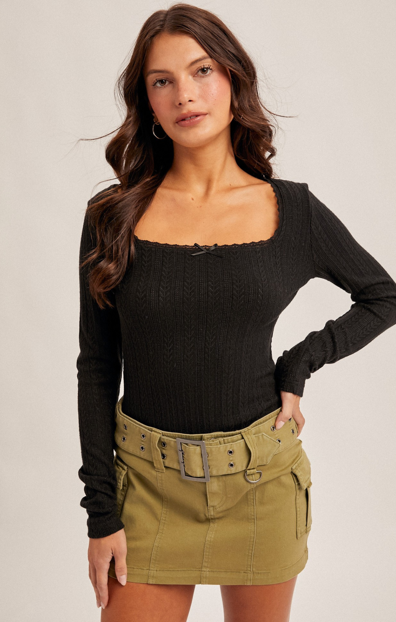 Woman wearing a black long-sleeve top and green skirt with a belt against a plain background