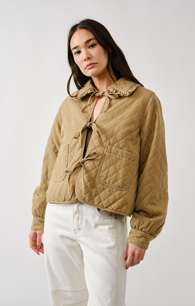 Woman wearing a beige quilted jacket and white pants on a plain background