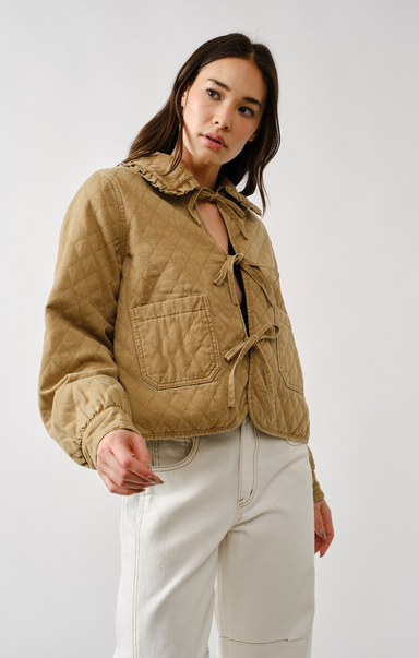 Woman wearing a beige quilted jacket and white pants against a plain background