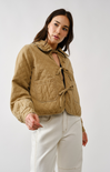 Woman wearing a beige quilted jacket and white pants against a plain background