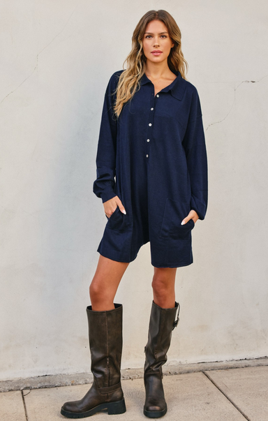Woman wearing a navy blue dress with long sleeves and knee-high brown boots against a white wall.