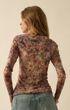 A woman shown from the back wearing a sheer, long-sleeve floral top with a muted pink, brown, and green floral pattern, paired with blue jeans, against a plain beige background.