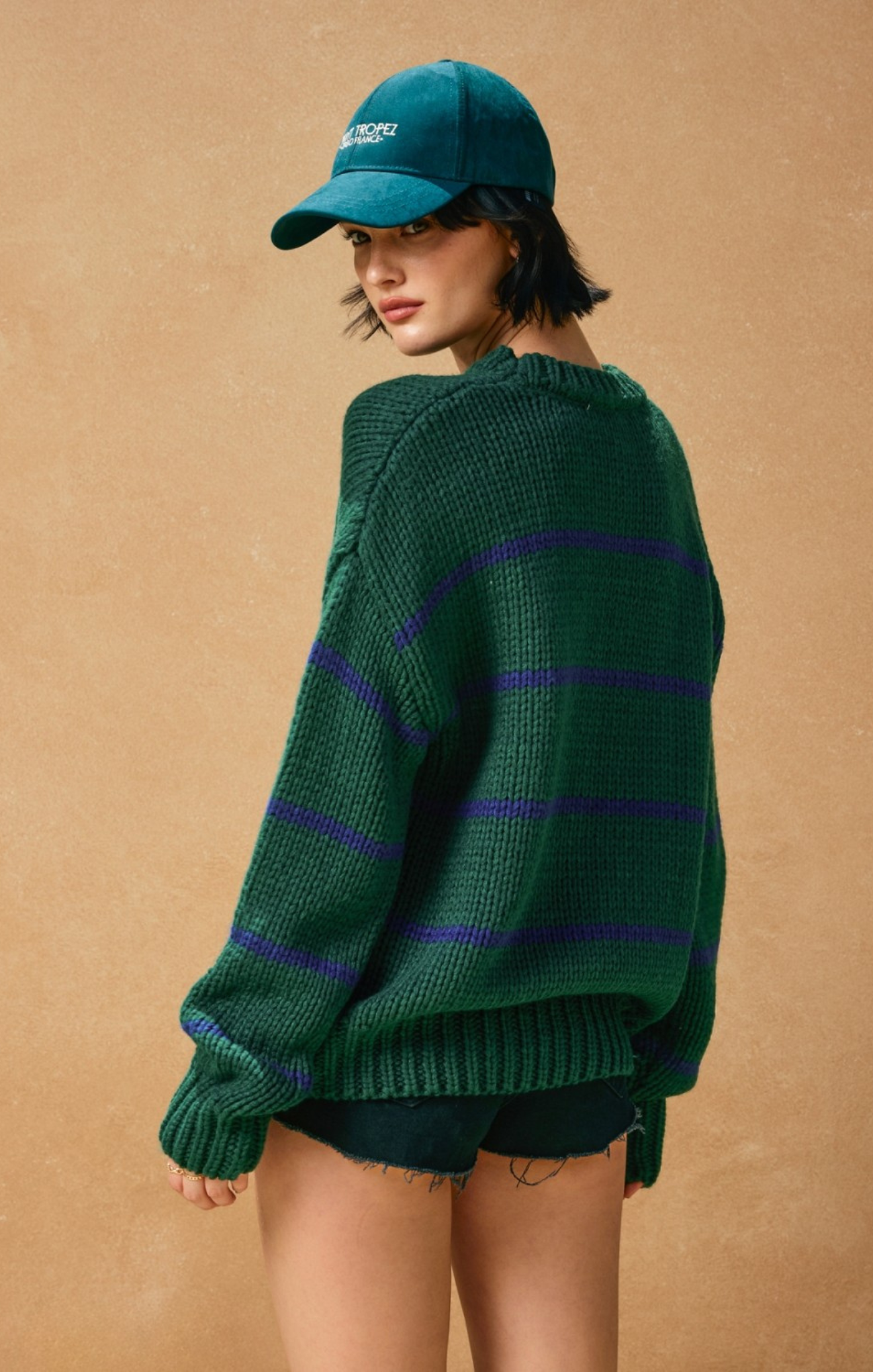 Person wearing a green sweater with purple stripes and a teal cap on a beige background