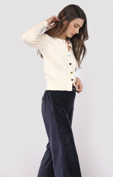 Woman wearing a cream cardigan with colorful buttons and navy pants on a white background
