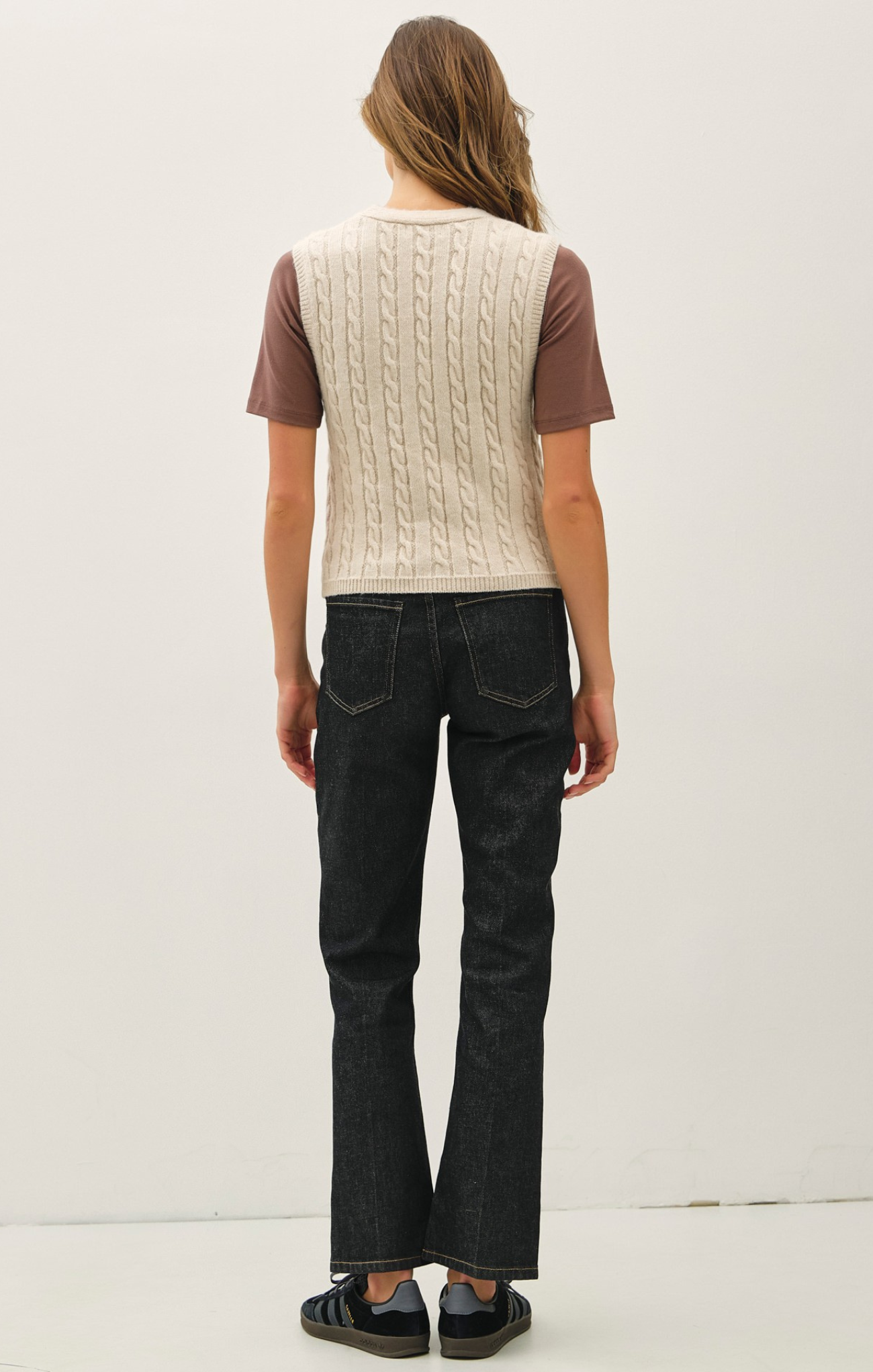 Person wearing a brown t-shirt with a beige cable knit vest and black jeans on a white background