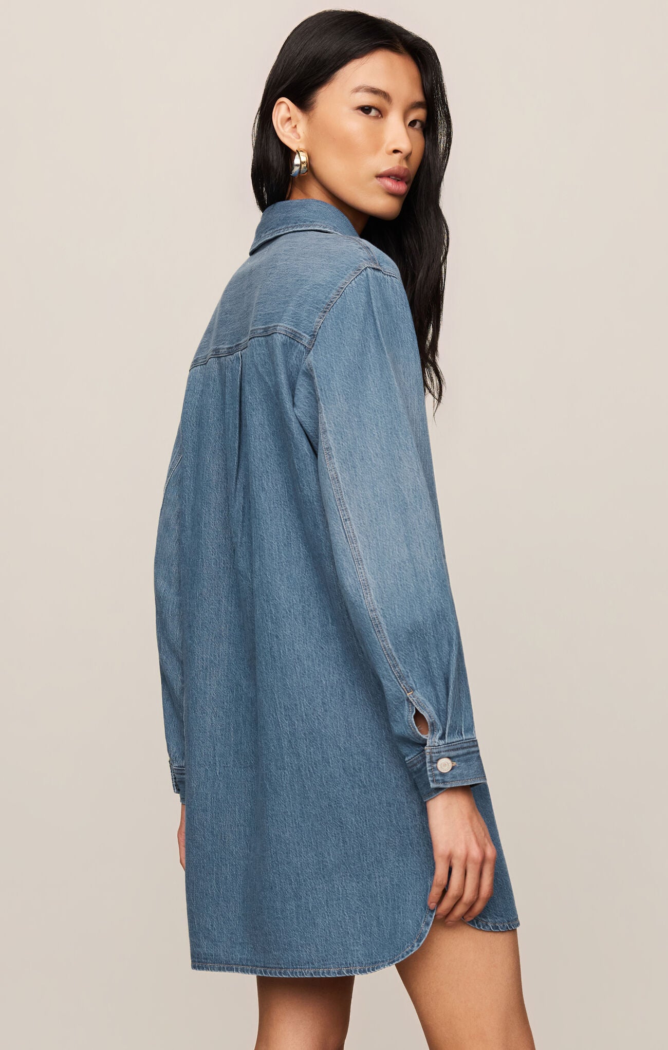 A woman modeling an oversized blue denim shirt dress, shown from a side-back angle against a plain beige studio background.