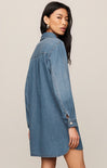 A woman modeling an oversized blue denim shirt dress, shown from a side-back angle against a plain beige studio background.