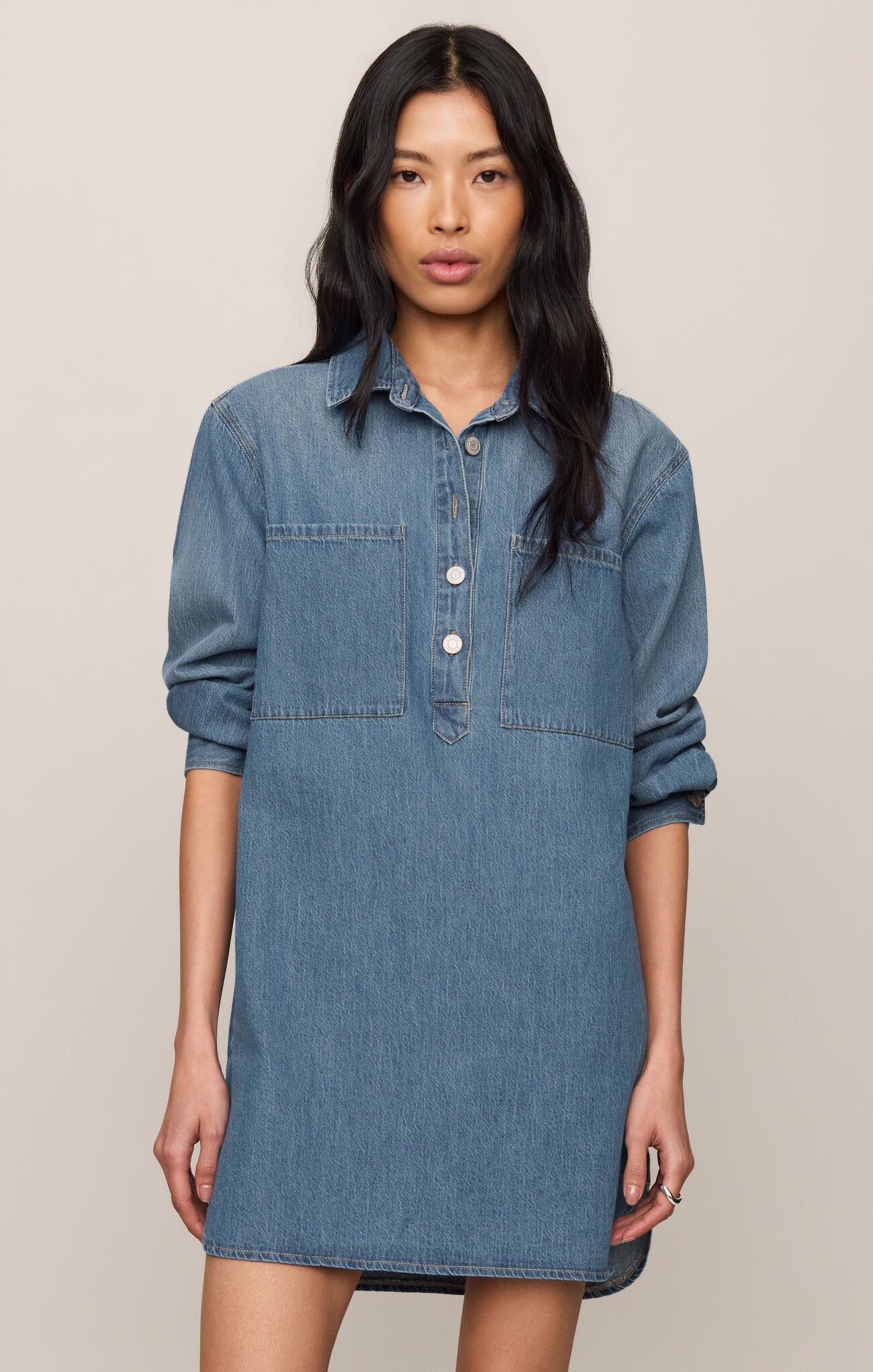 A woman modeling a medium-wash blue denim shirt dress with three-quarter sleeves, a buttoned collar, and two chest pockets, set against a plain beige background.