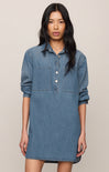 A woman modeling a medium-wash blue denim shirt dress with three-quarter sleeves, a buttoned collar, and two chest pockets, set against a plain beige background.