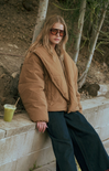 Woman in a beige puffer jacket and sunglasses leaning against a stone wall with a drink.