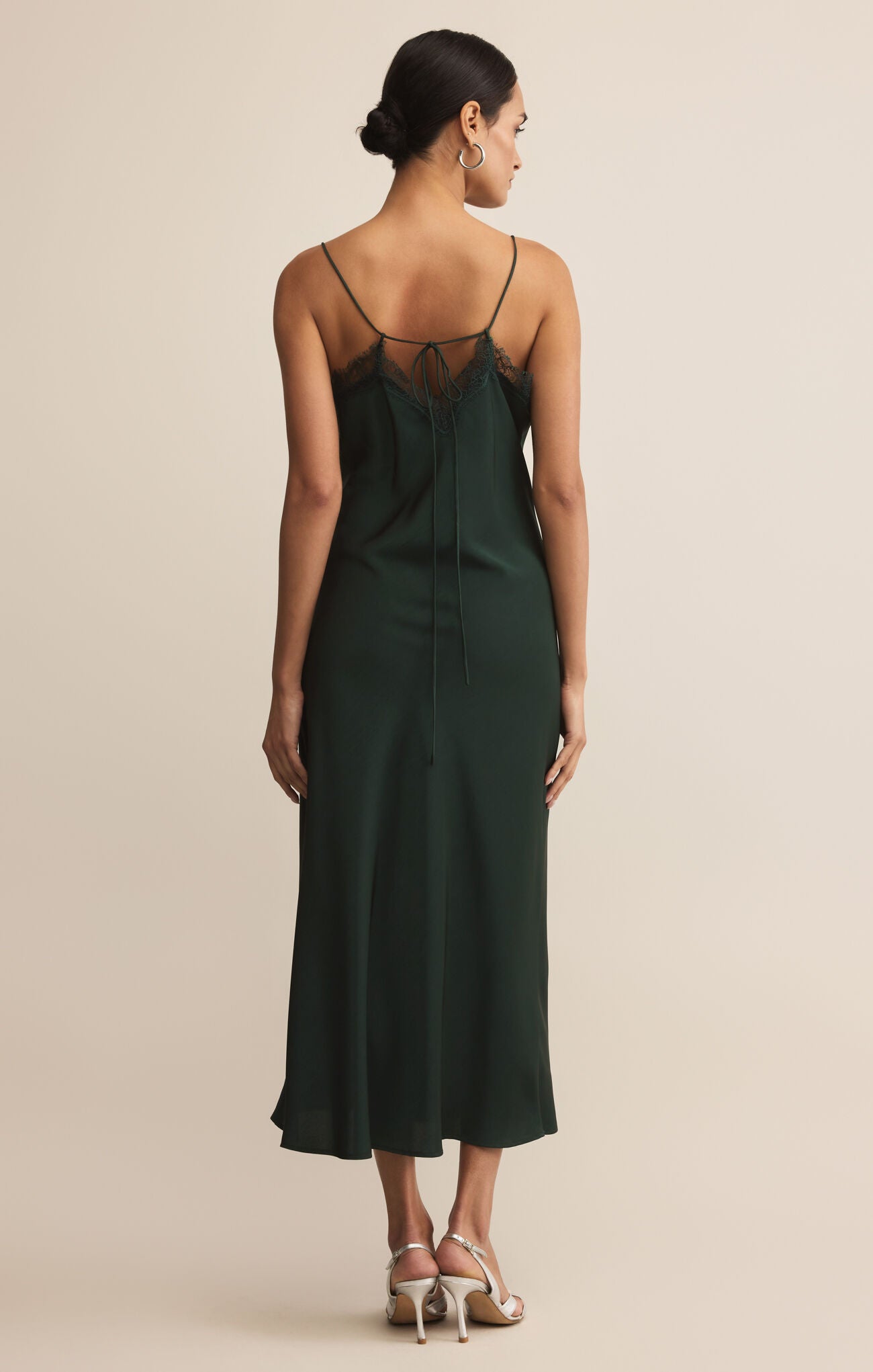 A woman wearing a dark green midi slip dress with lace trim and thin straps stands against a neutral background, paired with silver heels