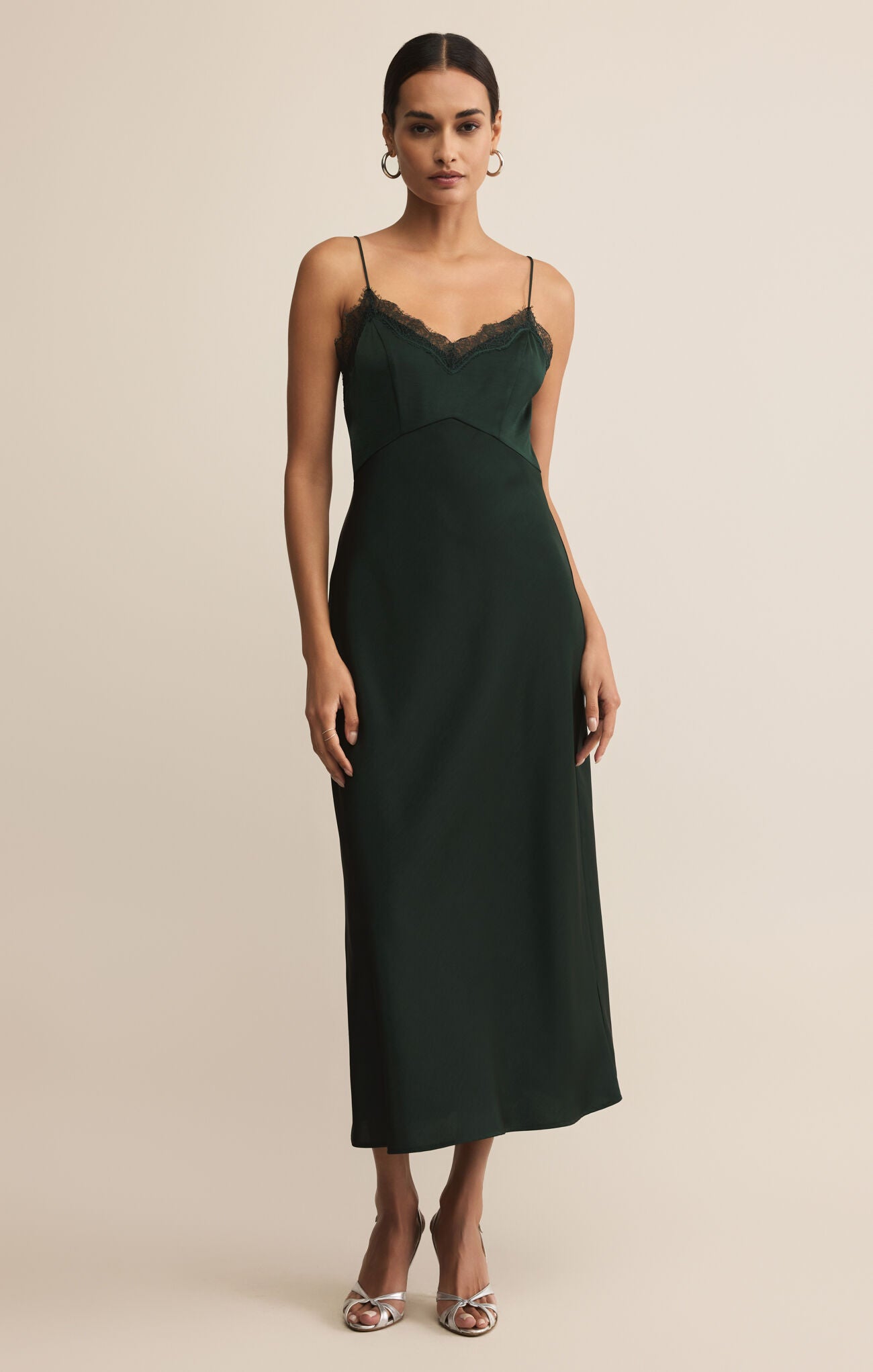 A woman wearing a dark green midi slip dress with lace trim and thin straps stands against a neutral background, paired with silver heels