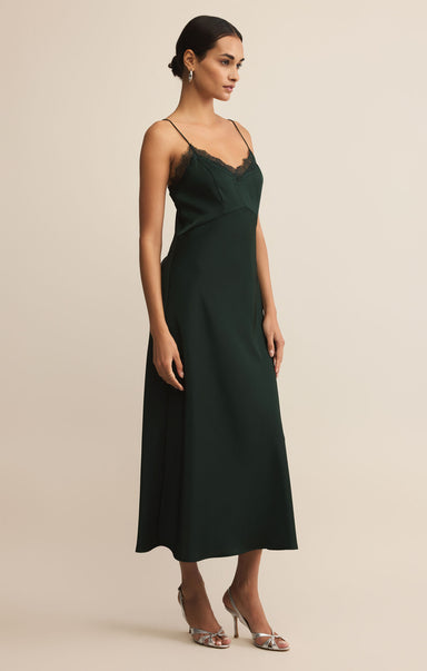 A woman wearing a dark green midi slip dress with lace trim and thin straps stands against a neutral background, paired with silver heels