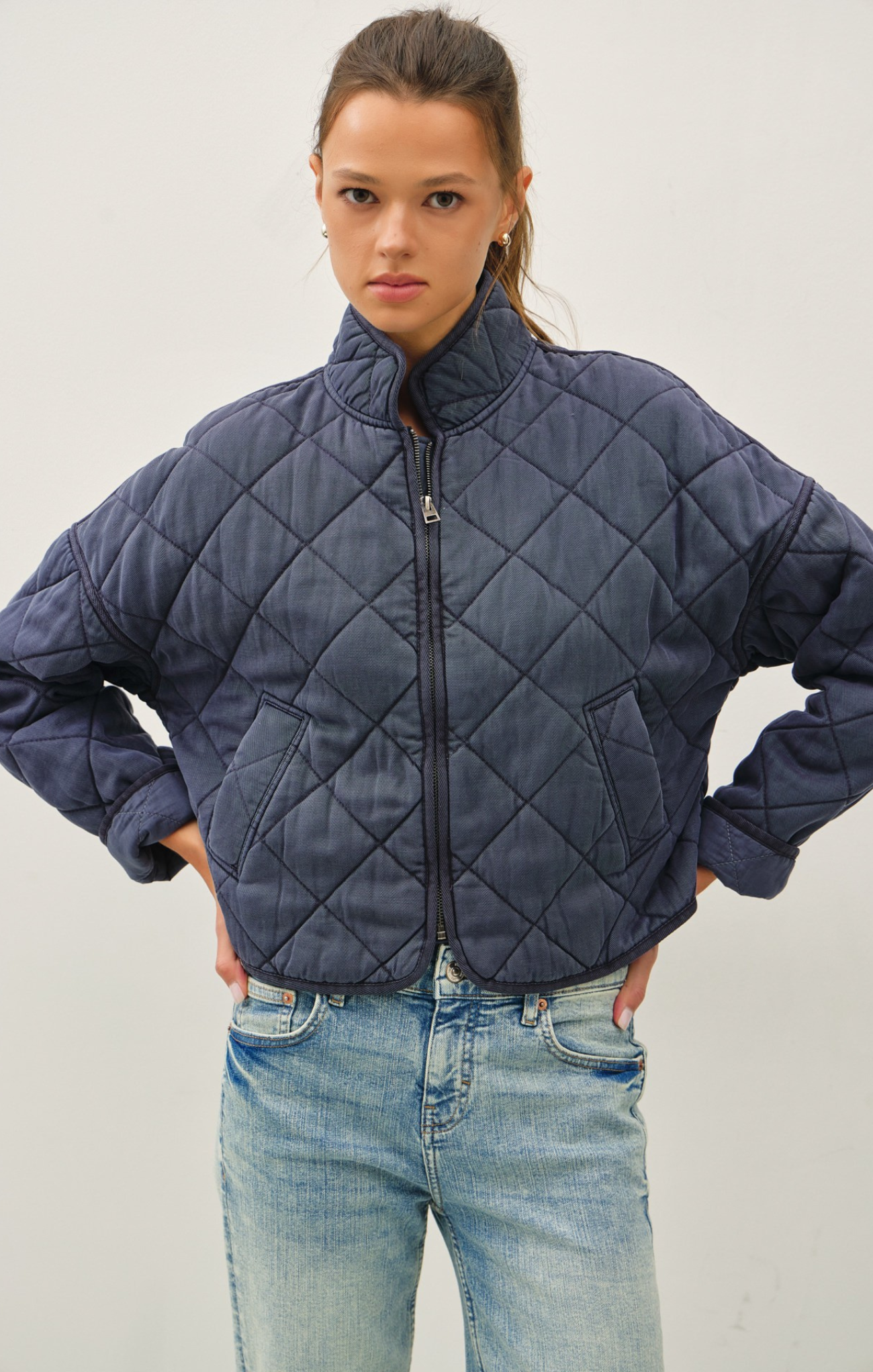 Woman wearing a charcoal-blue quilted cropped jacket with a high collar and front zipper, paired with light-wash jeans; standing with hands on hips against a plain light background.