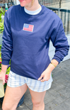 Person wearing a blue sweatshirt with an American flag patch and plaid shorts outdoors.