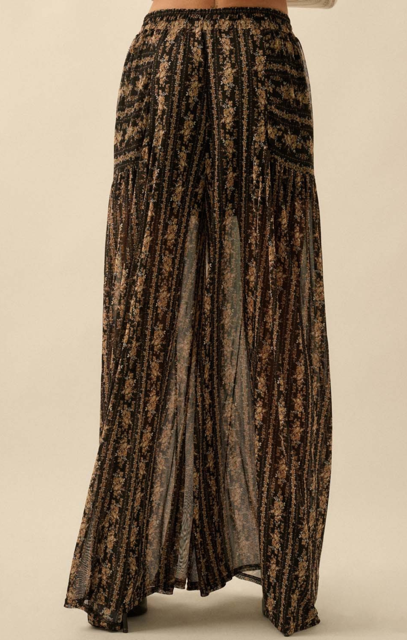 A close-up of a person wearing high-waisted, wide-leg sheer pants with a black base and gold floral pattern, featuring a flowy, lightweight silhouette.