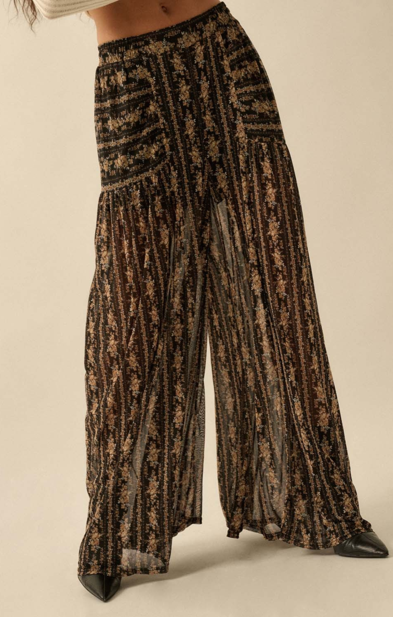 A close-up of a person wearing high-waisted, wide-leg sheer pants with a black base and gold floral pattern, featuring a flowy, lightweight silhouette.