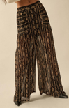 A close-up of a person wearing high-waisted, wide-leg sheer pants with a black base and gold floral pattern, featuring a flowy, lightweight silhouette.
