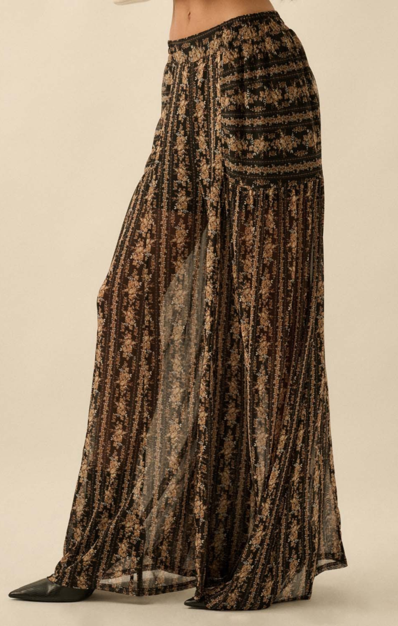 A close-up of a person wearing high-waisted, wide-leg sheer pants with a black base and gold floral pattern, featuring a flowy, lightweight silhouette.