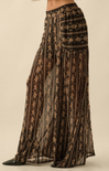 A close-up of a person wearing high-waisted, wide-leg sheer pants with a black base and gold floral pattern, featuring a flowy, lightweight silhouette.
