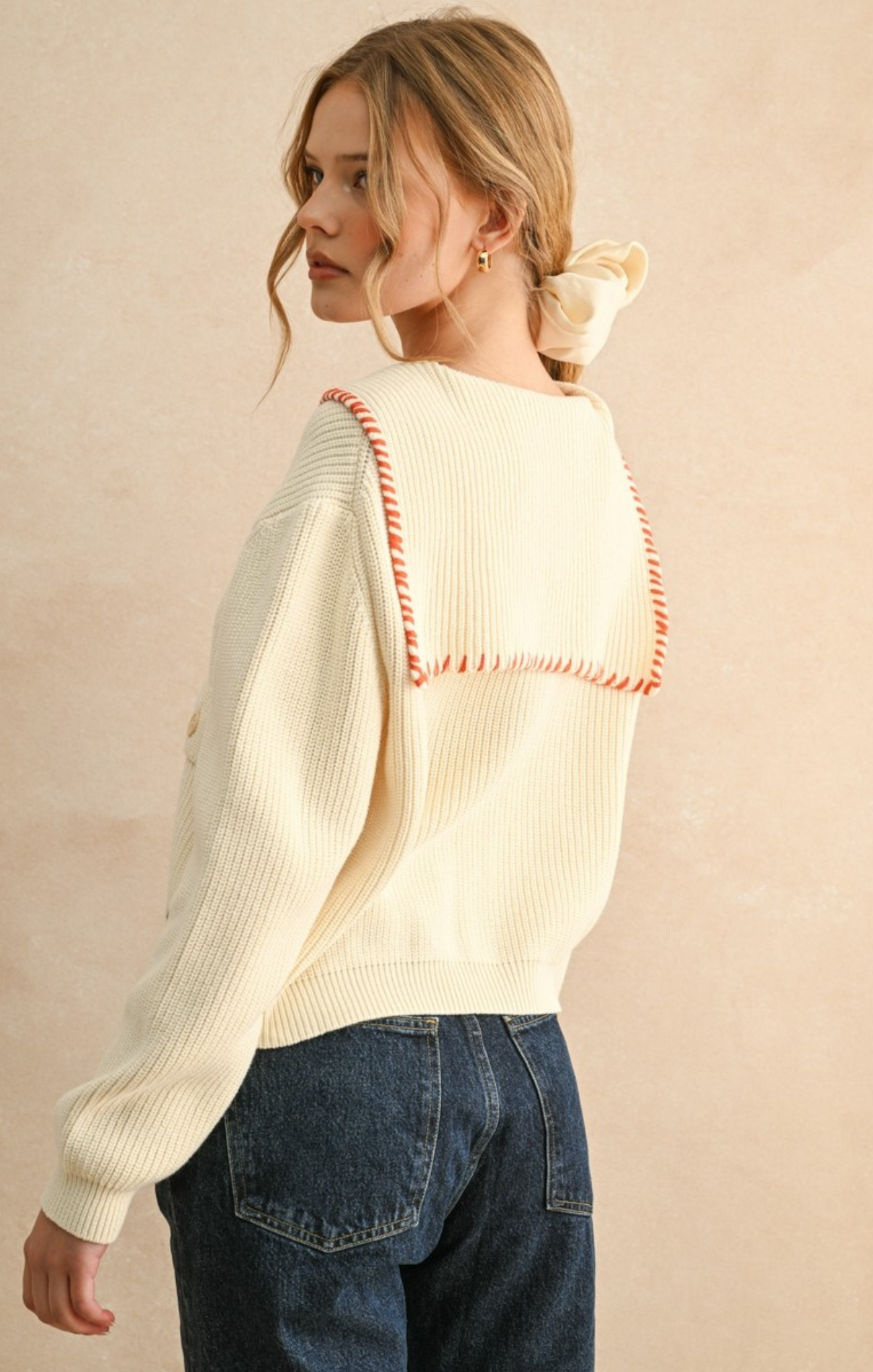 Woman wearing a cream sweater with red accents and blue jeans against a beige background