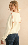 Woman wearing a cream sweater with red accents and blue jeans against a beige background