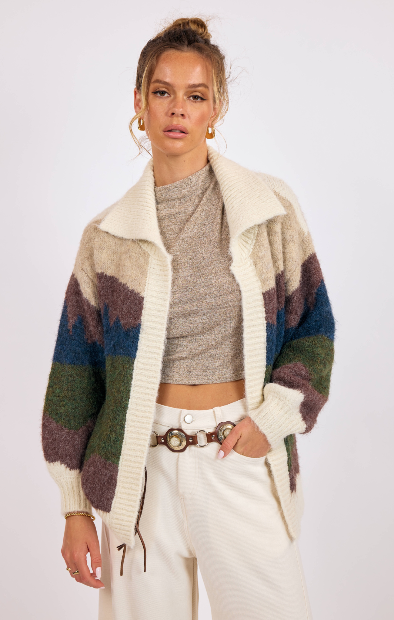 Woman wearing a patterned cardigan over a crop top with white pants on a plain background