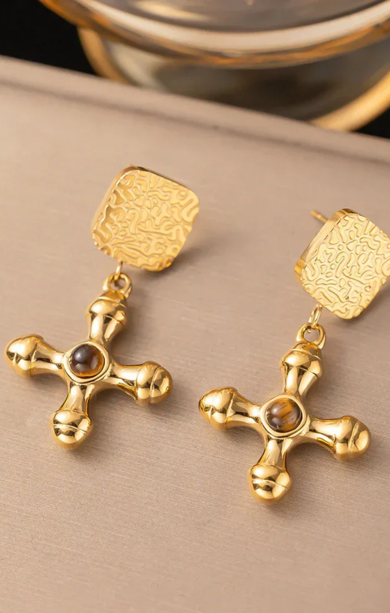 Gold cross earrings with embedded stones on a beige surface