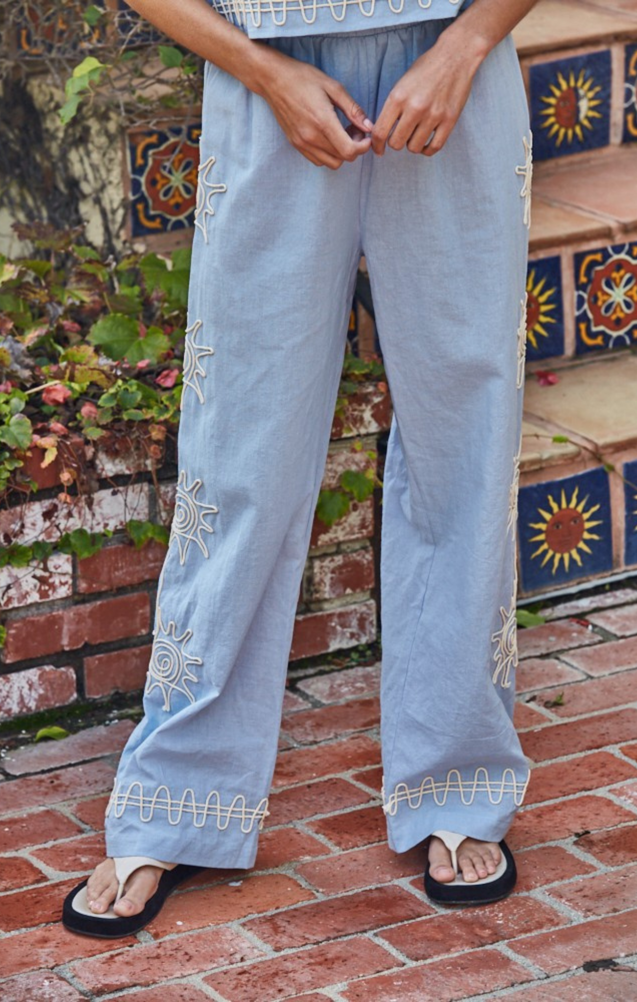Light blue pants with embroidery on a person standing on a brick floor with decorative tiles in the background.