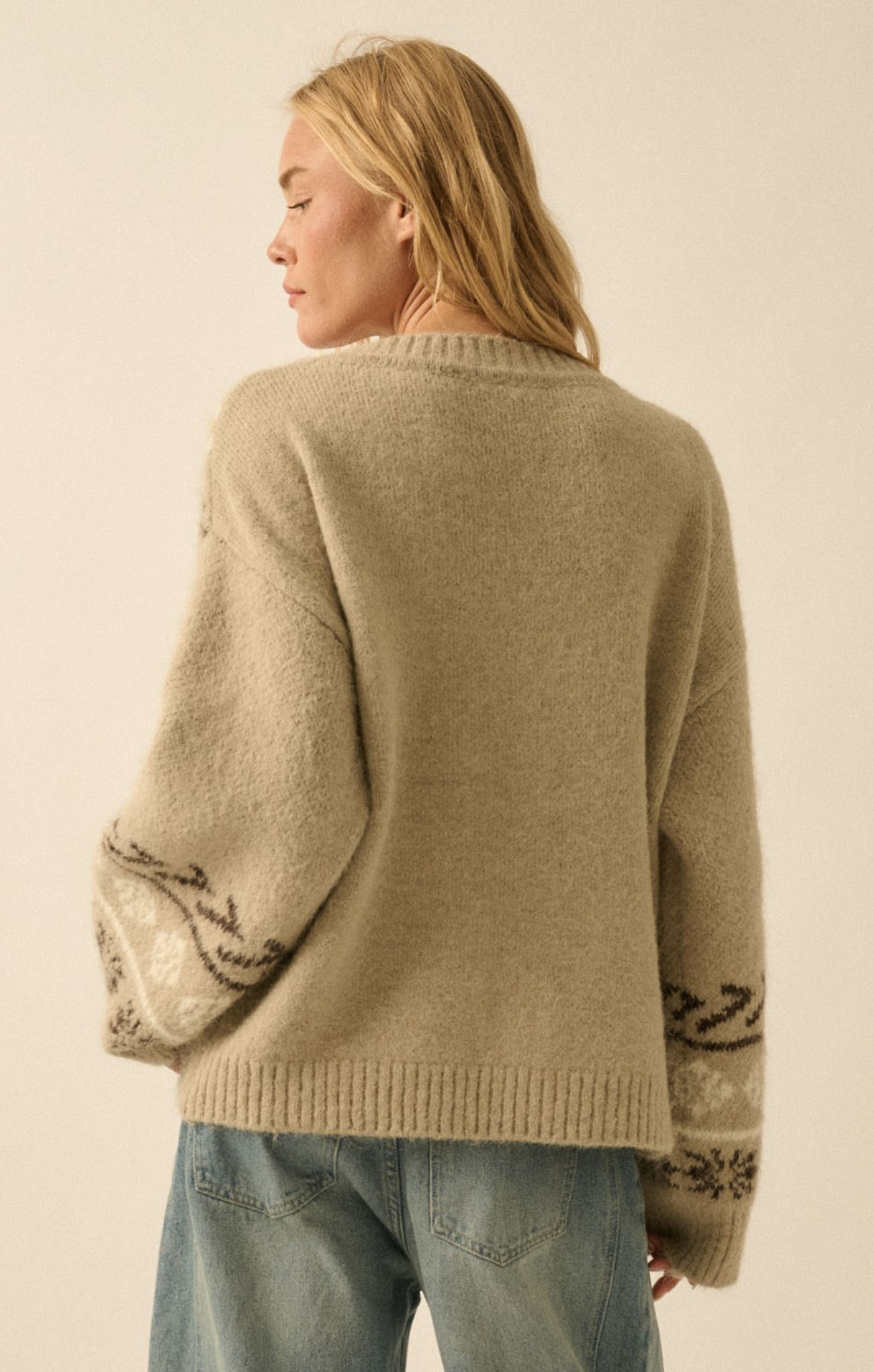 Person wearing a beige sweater with patterned cuffs and hem on a plain background