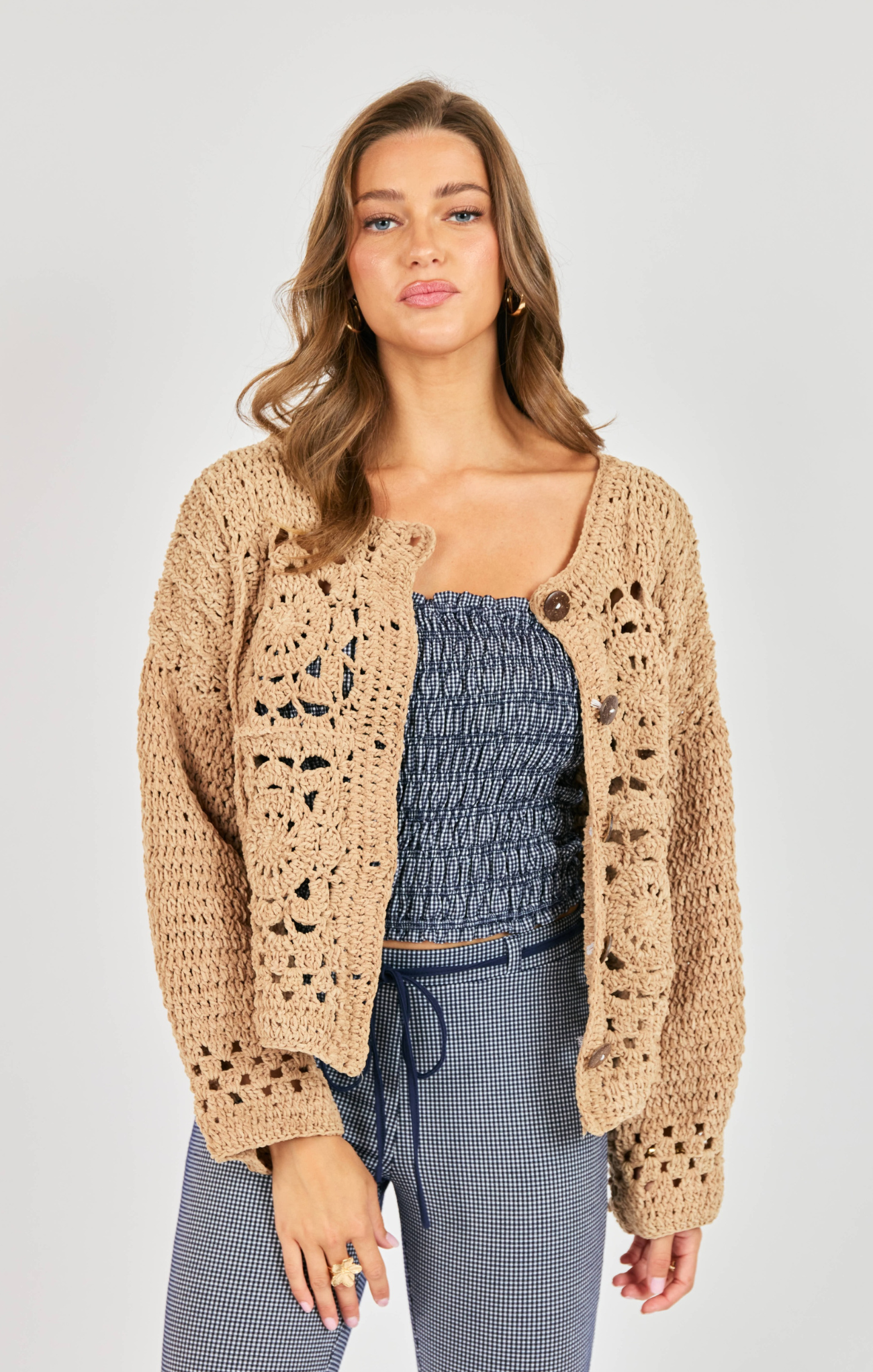 Woman wearing a beige crochet cardigan over a blue top and pants on a white background