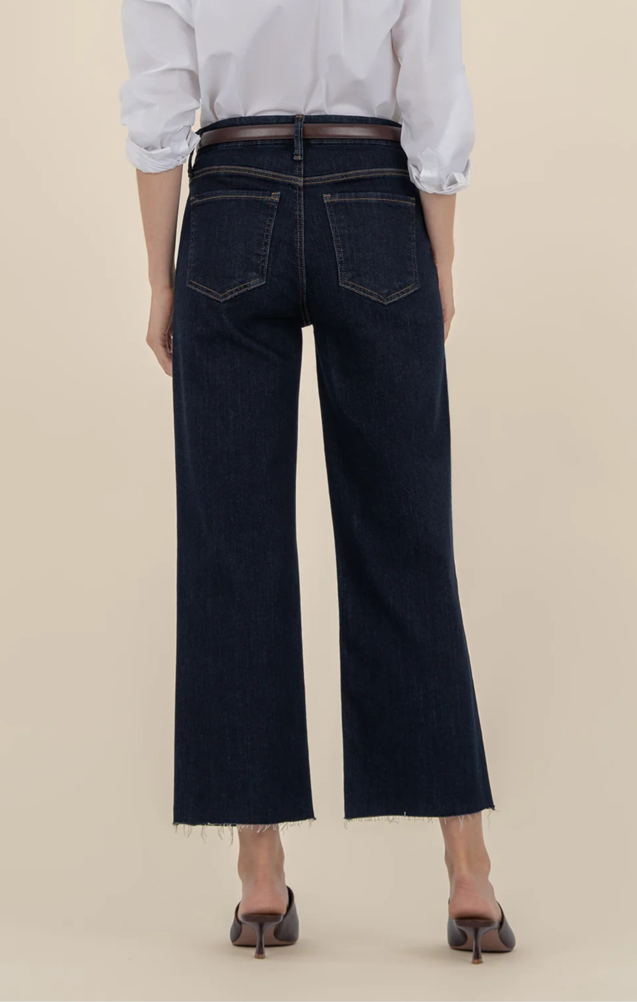 Person wearing dark-wash wide-leg jeans with front patch pockets and a raw hem, styled with a white blouse, brown belt, and brown slide sandals, shown from shoulders to feet against a neutral background.