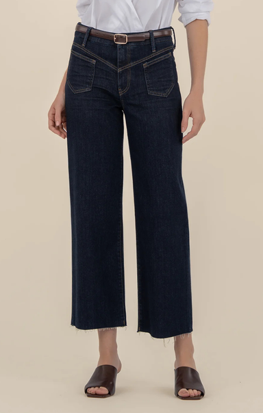 Person wearing dark-wash wide-leg jeans with front patch pockets and a raw hem, styled with a white blouse, brown belt, and brown slide sandals, shown from shoulders to feet against a neutral background.