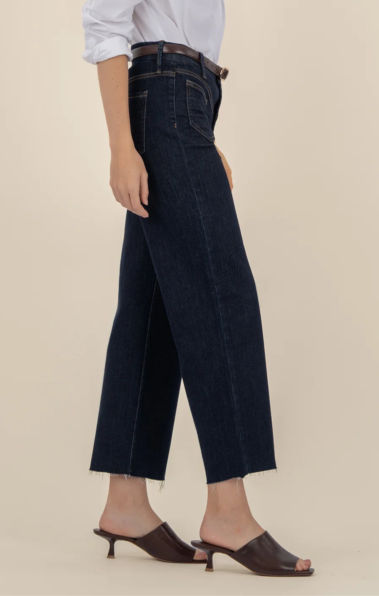 Person wearing dark-wash wide-leg jeans with front patch pockets and a raw hem, styled with a white blouse, brown belt, and brown slide sandals, shown from shoulders to feet against a neutral background.