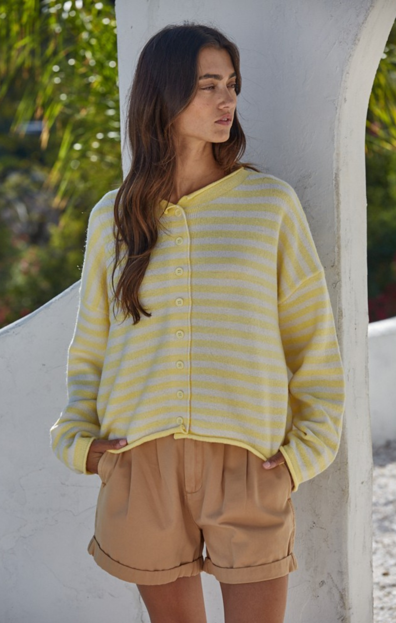 Woman wearing a yellow striped cardigan and beige shorts standing against a white wall with greenery in the background.