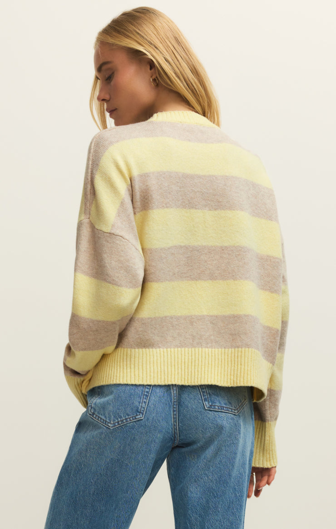 Woman wearing a yellow and gray striped sweater with blue jeans on a plain background
