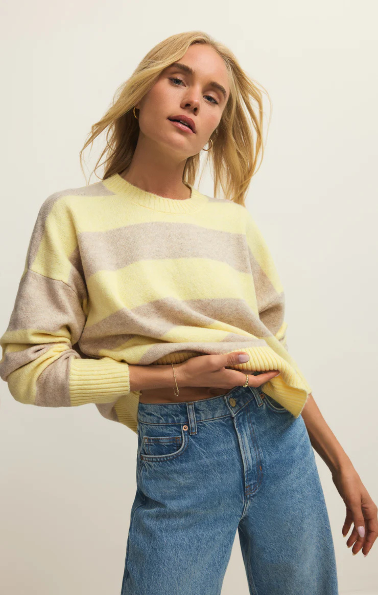 Woman wearing a yellow and beige striped sweater with blue jeans against a plain background