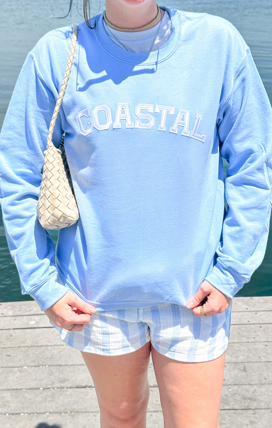Person wearing a light blue sweatshirt with 'COASTAL' text, striped shorts, and holding a woven bag.