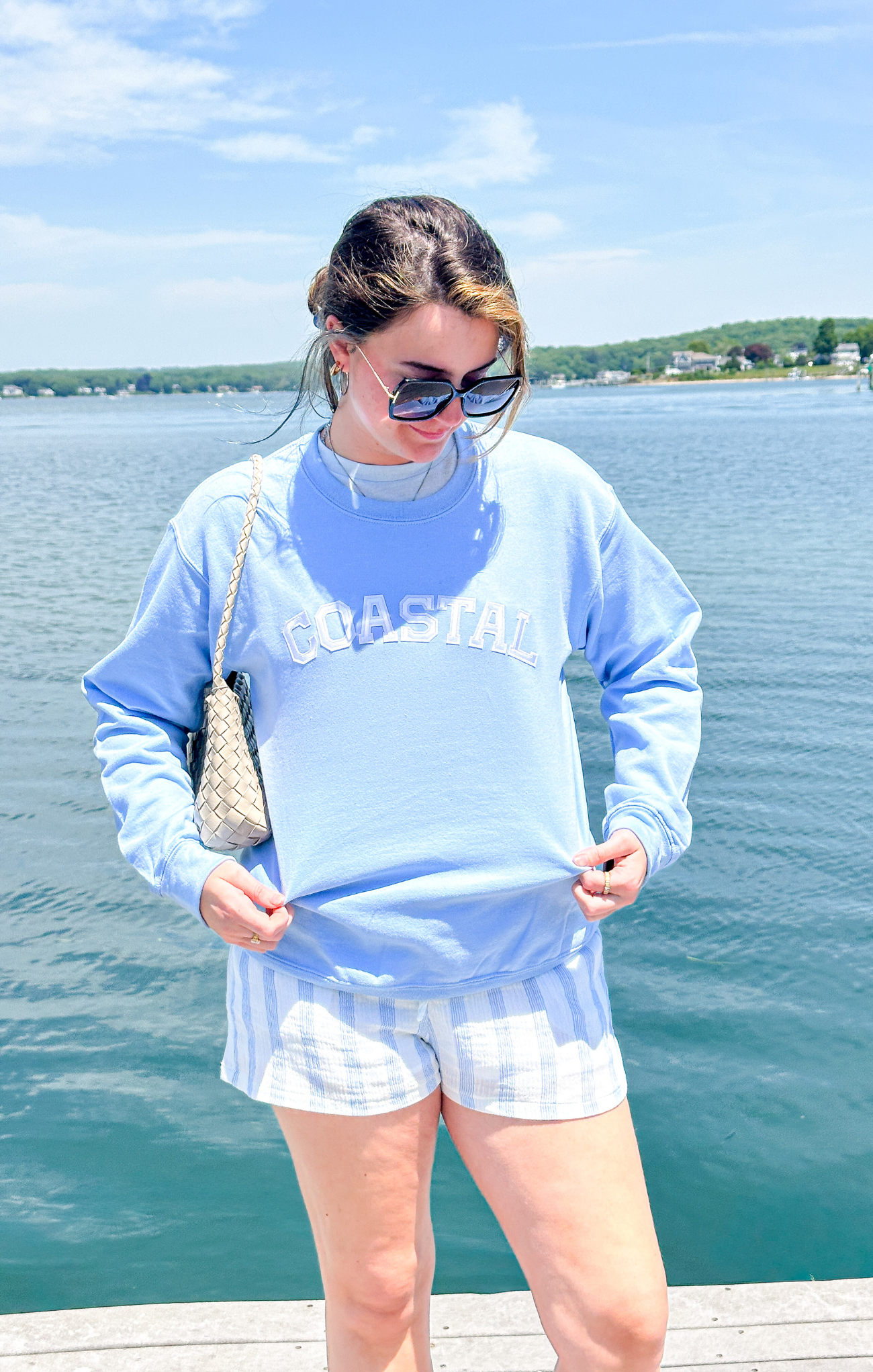 Woman wearing a light blue sweatshirt with text, white shorts, and sunglasses by a lake.