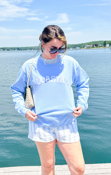 Woman wearing a light blue sweatshirt with text, white shorts, and sunglasses by a lake.