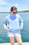 Woman wearing a light blue sweatshirt with text, white shorts, and sunglasses by a lake.