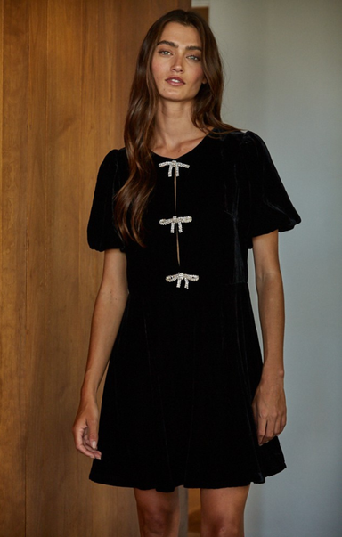 Woman wearing a black dress with white bow details against a wooden wall.