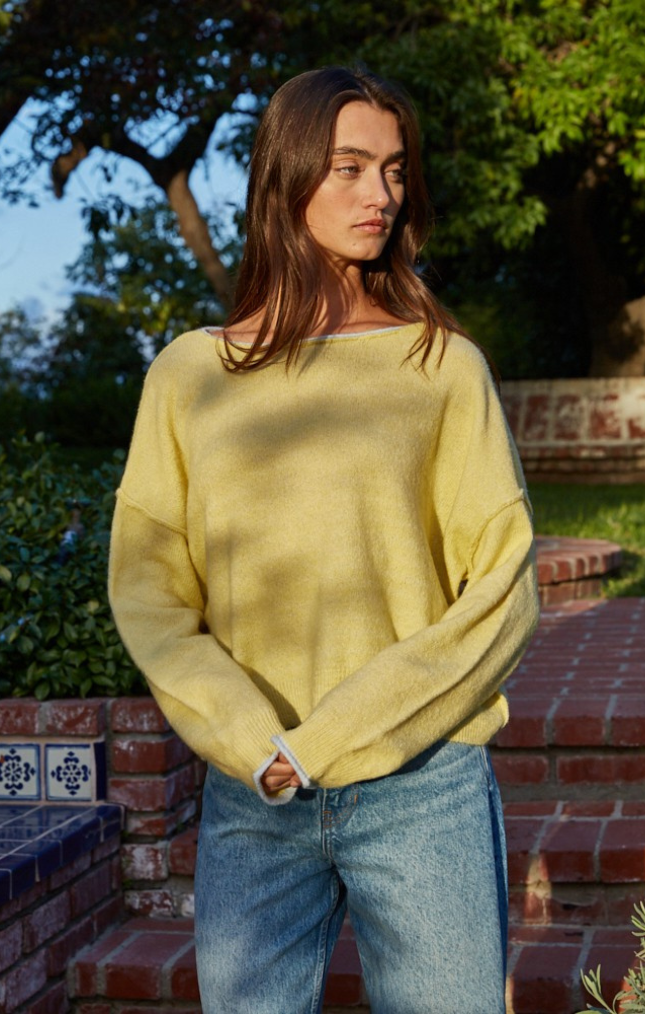 Person wearing a yellow sweater and blue jeans standing outdoors with greenery in the background