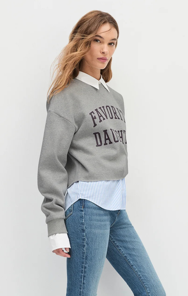 Woman wearing a gray sweatshirt with text, white shirt, and blue jeans on a plain background