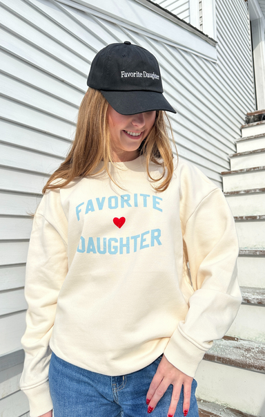 Person wearing a cream sweatshirt with 'Favorite Daughter' text and a black cap outdoors.