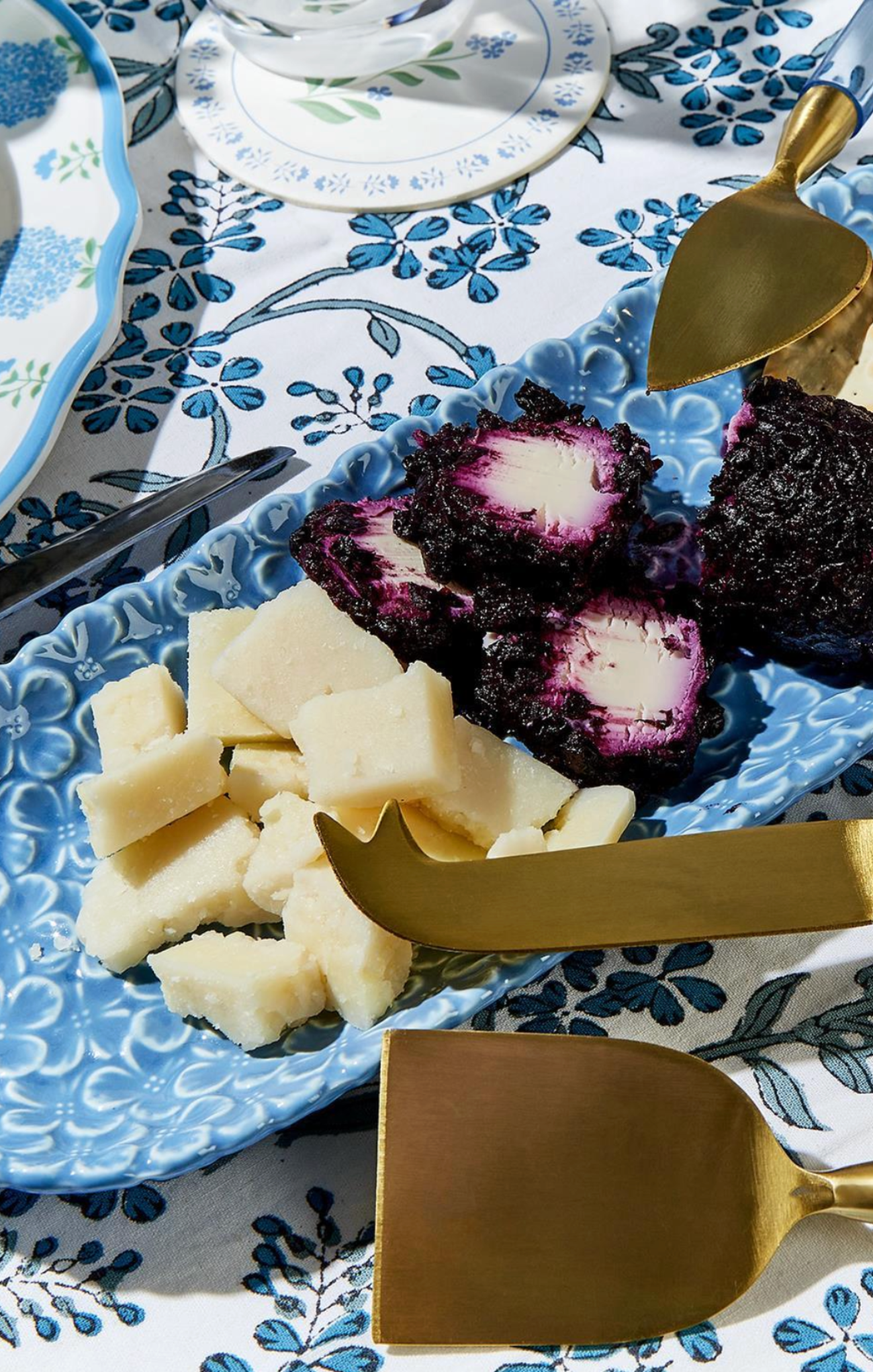Cheese and purple fruit on a blue patterned plate with gold cheese knives.