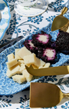 Cheese and purple fruit on a blue patterned plate with gold cheese knives.