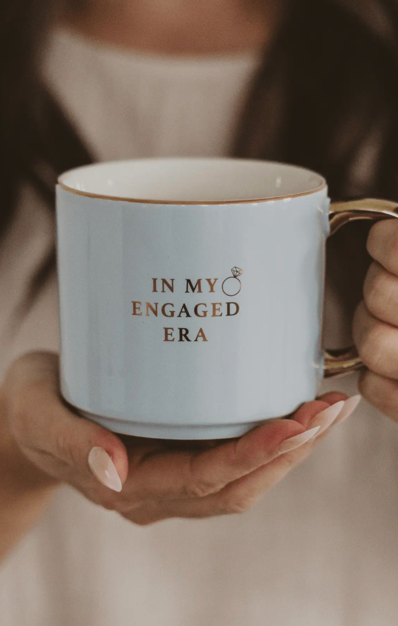 Close-up of hands holding a light blue ceramic mug with a gold handle and rim, featuring the phrase “In My Engaged Era” in gold lettering with a small engagement ring icon above the text.