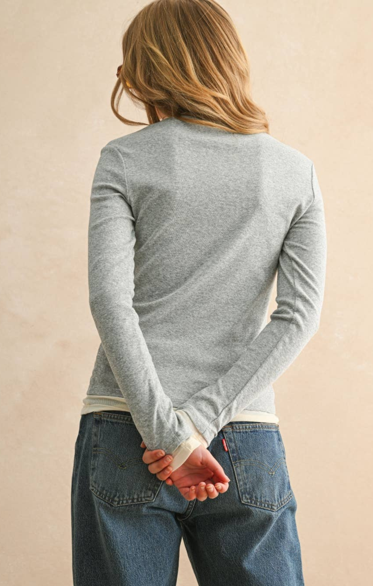 Person wearing a gray long-sleeve shirt and blue jeans against a beige background
