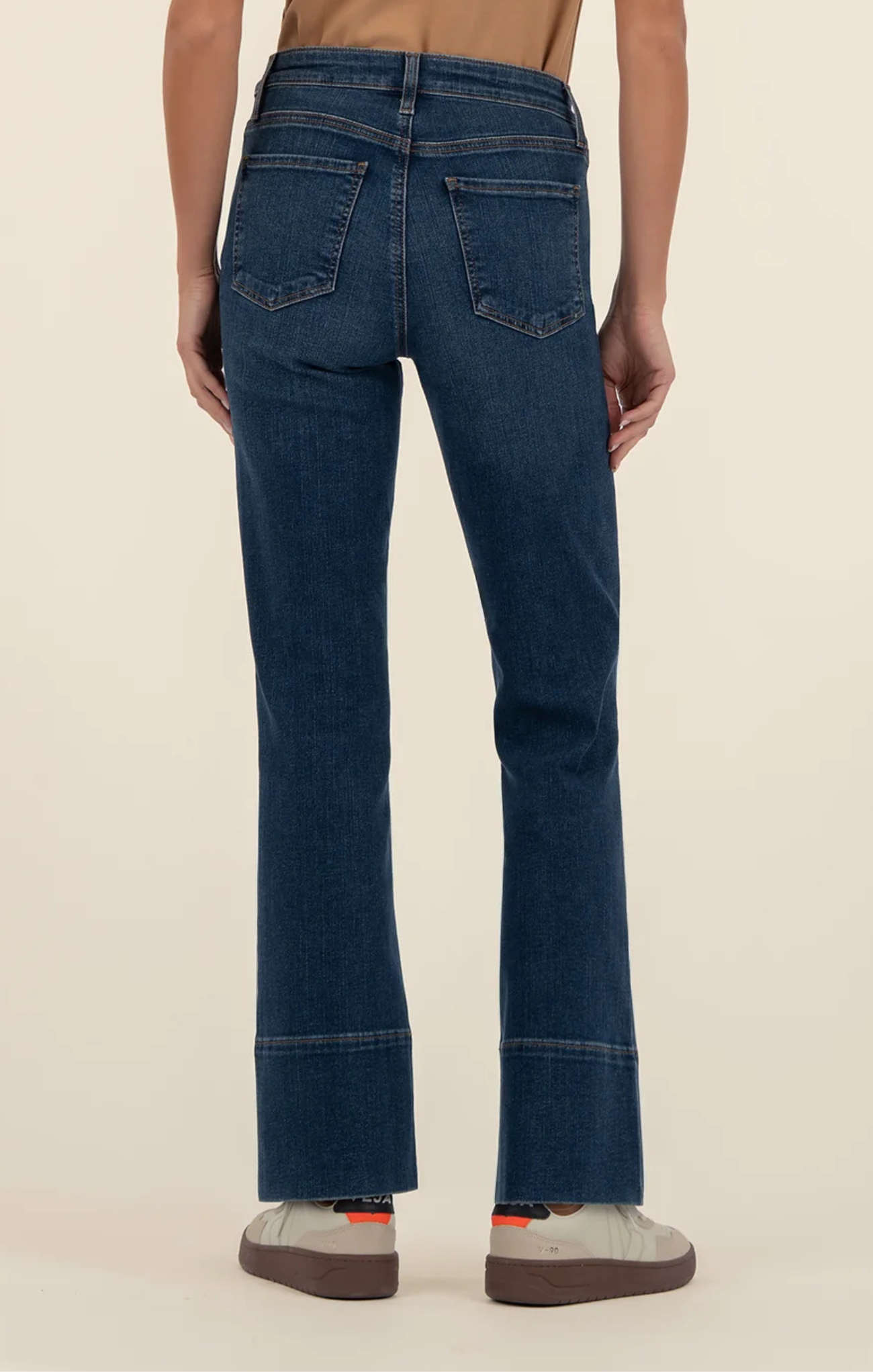 Person wearing dark-wash high-rise flare jeans with front seam detailing, paired with a tan top and light gray sneakers, shown from shoulders to feet against a neutral background.